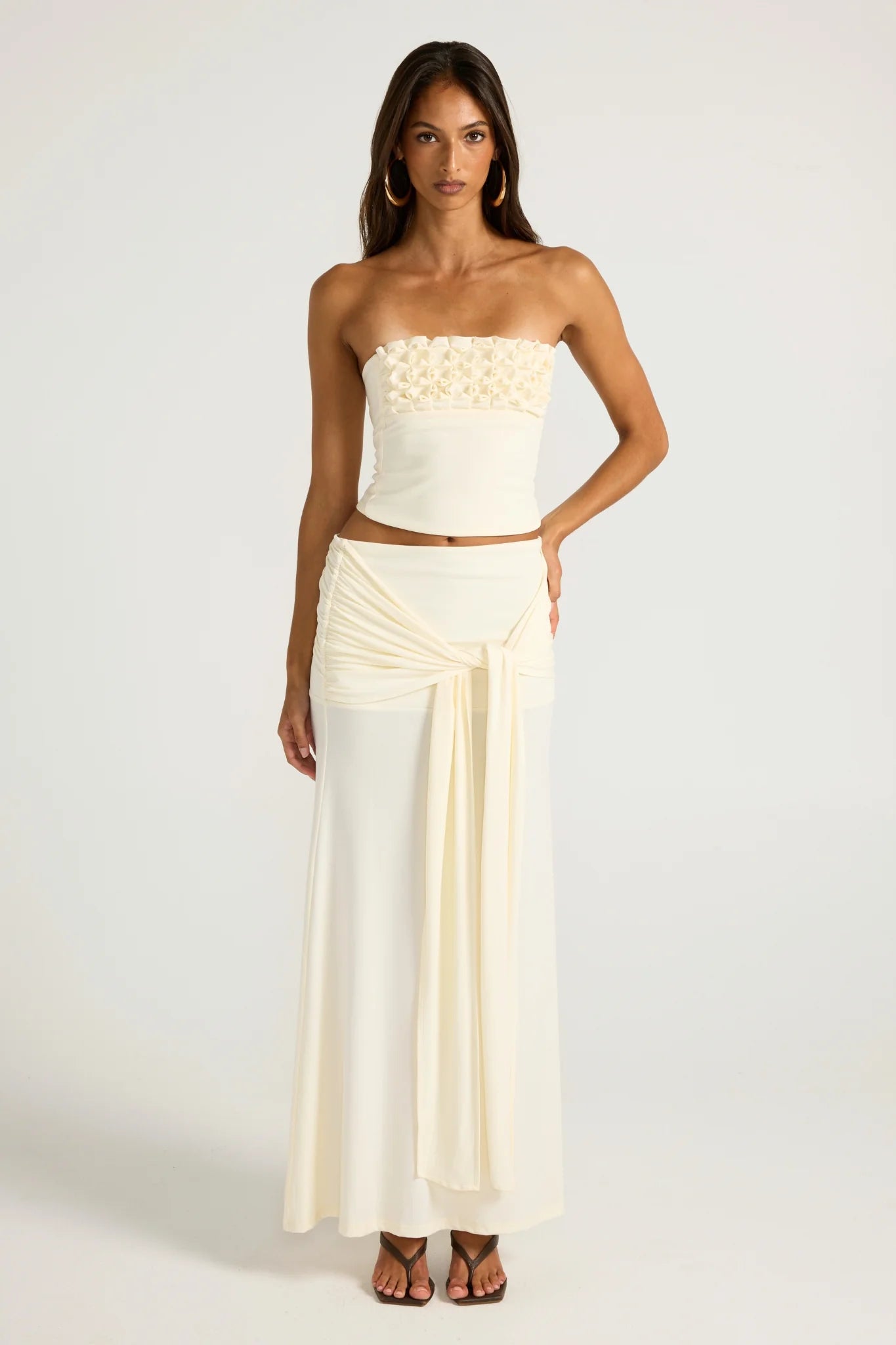 Ance Gria Jude Set in Ivory for hire. Strapless bandeau top with 3D floral smocking and matching low-rise sheer maxi skirt with long tie detail.