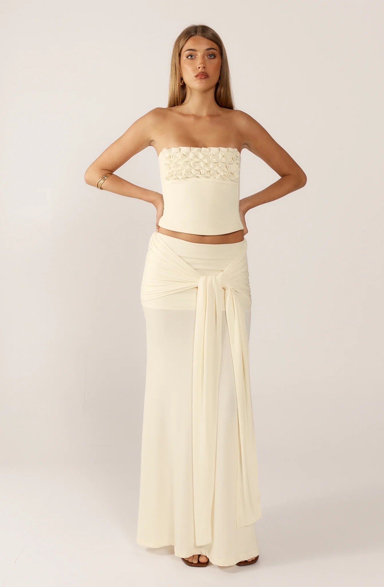 Ance Gria Jude Set in Ivory for hire. Strapless bandeau top with 3D floral smocking and matching low-rise sheer maxi skirt with long tie detail.